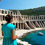 A person in modest, appropriate attire suitable for exploring historical sites, stands thoughtfully amidst ancient Roman and Greek ruins on the Turkish coast. Majestic columns and an ancient amphitheater are visible, with crystal-clear turquoise Mediterranean waters in the background under a bright, sunny sky. The atmosphere is serene and historical. Safe for work, appropriate content, fully clothed, professional, perfect anatomy, correct proportions, natural pose, well-formed hands, proper finger count, natural body proportions, professional photography, high quality.